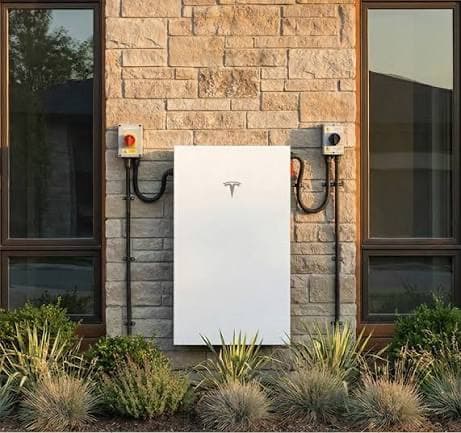 A Tesla Powerwall mounted on the exterior stone wall of a house, with conduit running to disconnects either side